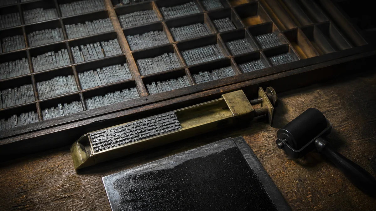 A letterpress tray of metal type sorts with a composing stick and ink roller.