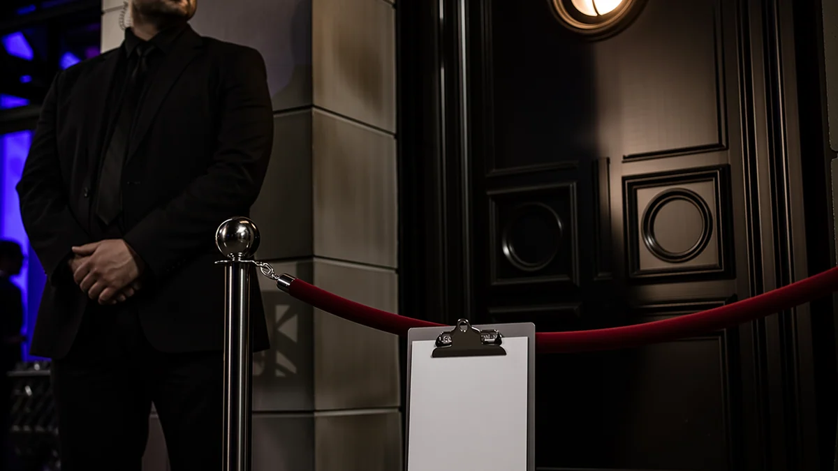A velvet rope and a clipboard guest list at a dark door with a small lamp, door-policy mood.
